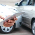 Seguros para carros em pernambuco representado por uma batida de carro no fundo, e com um tablet na mão fazendo anotações.