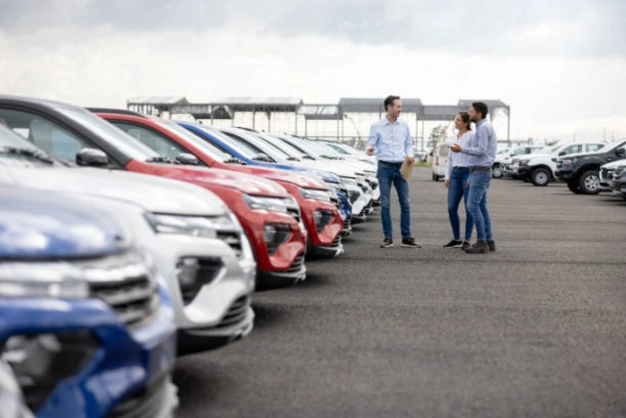 Vendedor fala com clientes em uma loja de seminovos, em frente a uma fileira de carros à venda. Como consultar tabela FIPE pela placa?