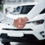 Duas pessoas dão as mãos em frente a um carro, representando 10 carros mais vendidos no Brasil.