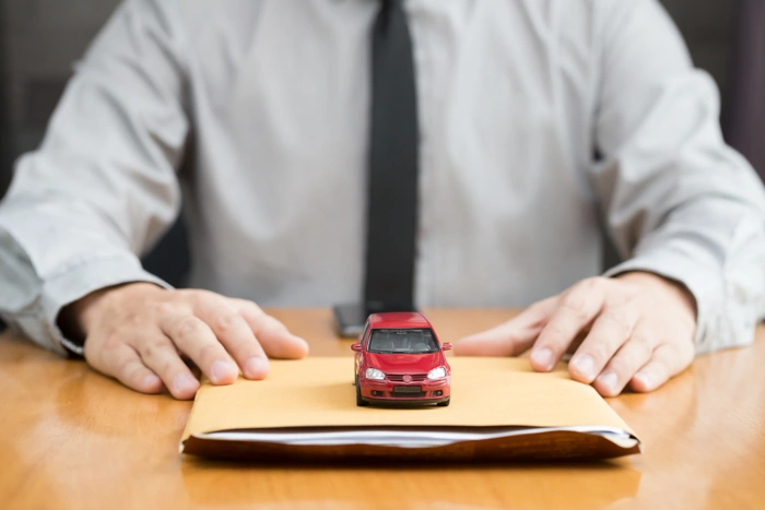 Homem sentado, a sua frente há um envelope com um carro em miniatura vermelho sobre ele. O homem está com as mãos sobre o envelope. Explicando se a seguradora star é boa.