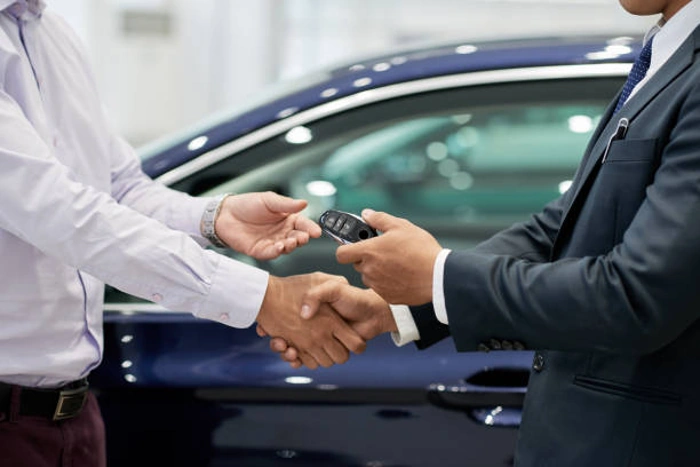 Vendedor cumprimenta comprador de carro, enquanto lhe entrega a chave do veículo em mãos. Explicando qual a diferença entre seguro e proteção veicular.