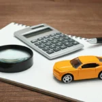 Há na imagem uma miniatura de carro amarelo, uma calculadora e uma lupa sobre uma folha branca. Falando seguro de carro barato e confiável.