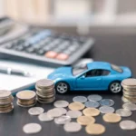 Pilhas de moedas, um prancheta, uma calculadora, uma caneta e uma miniatura de carro na cor azul sobre uma mesa. Falando sobre carros com o seguro mais caro.