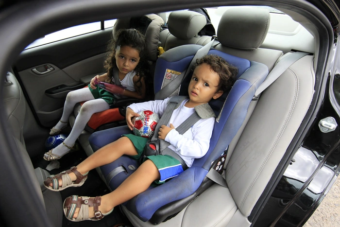 Dupla de irmãos sentados em cadeirinhas em banco de carro. Falando sobre qual é a cadeirinha certa para cada idade.