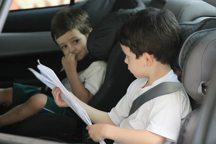 Dois meninos bem firmados no banco de trás de um carro. Um deles tem um papel nas mãos. Falando sobre qual é a cadeirinha certa para cada idade.